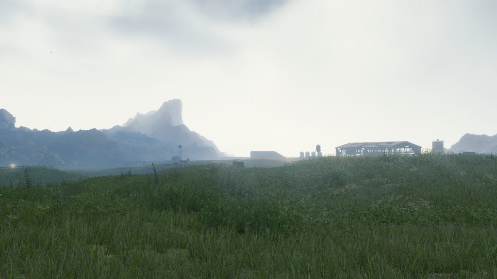 An image of a defunct barn and silos in the distance across a grassy expanse with a grey sky and mountains in the distance.